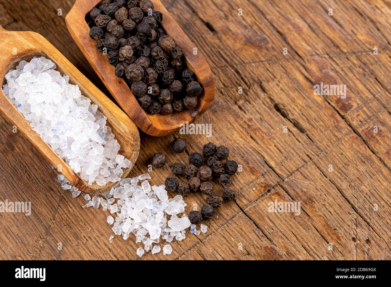 Nahaufnahme von grobem Kristallsalz und ganzen schwarzen Pfefferkörnern auf kleinen Gewürzschaufeln aus olivem Holz auf rustikalem Holzhintergrund Stockfoto