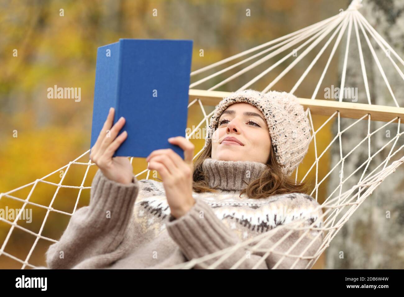 Entspannte und zufriedene Frau, die Papierbuch auf einer Hängematte liest In einem Wald im Herbst Stockfoto