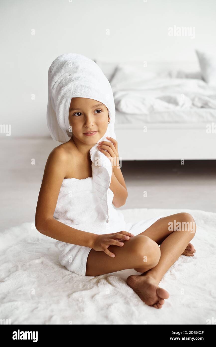 Morgendliche Routine. Kleines Mädchen in ein weißes Handtuch gewickelt nach einer Dusche sitzt auf dem Teppich auf dem warmen Boden in einem hellen Raum. Beauty, Spa und Hygiene Co Stockfoto