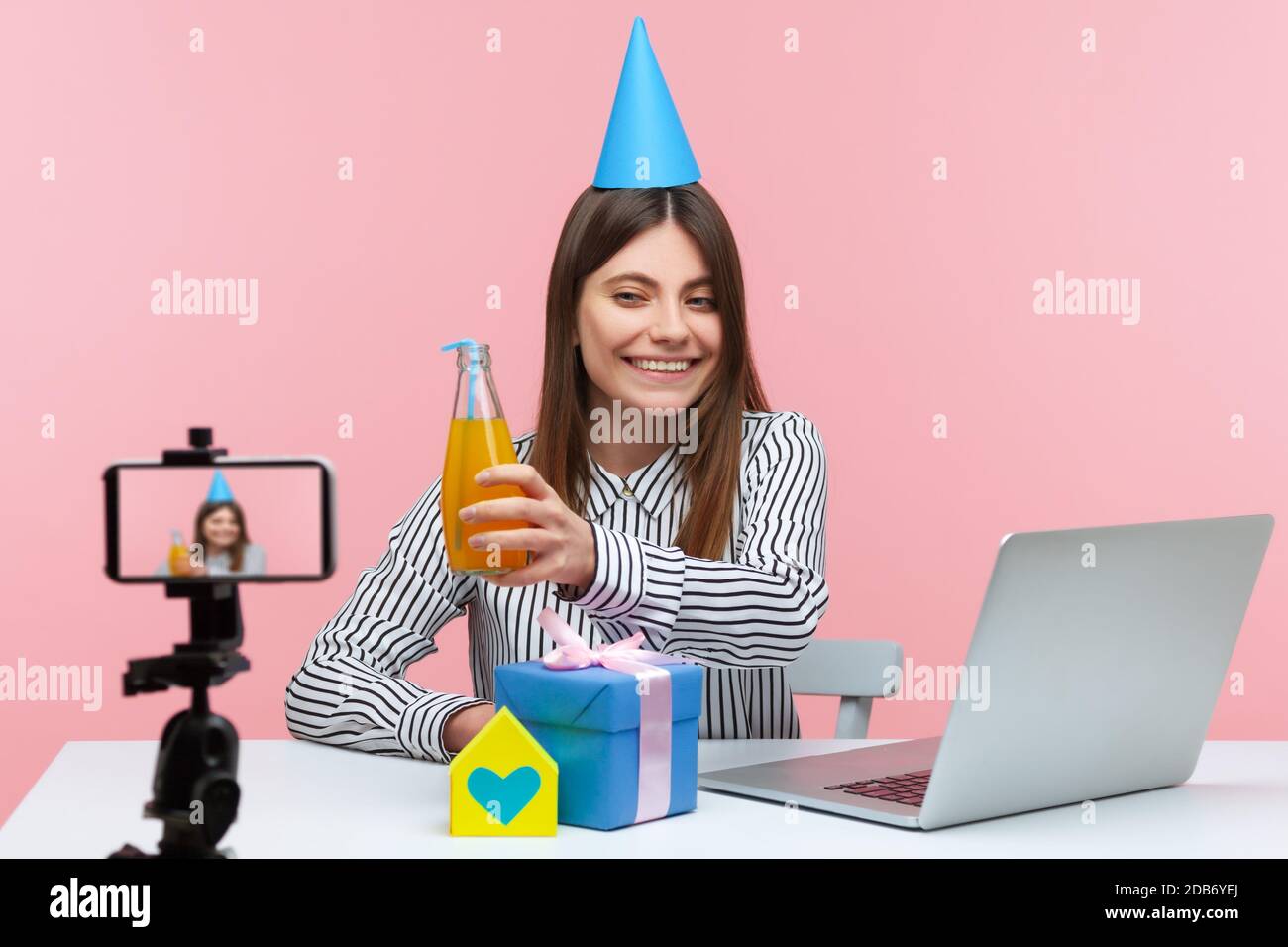 Positive lächelnde Brünette Frau sitzt vor der Kamera in Kegelhut Geburtstag feiern online, Geschenkbox und Papierhaus auf dem Schreibtisch in der Nähe Laptop. Stockfoto