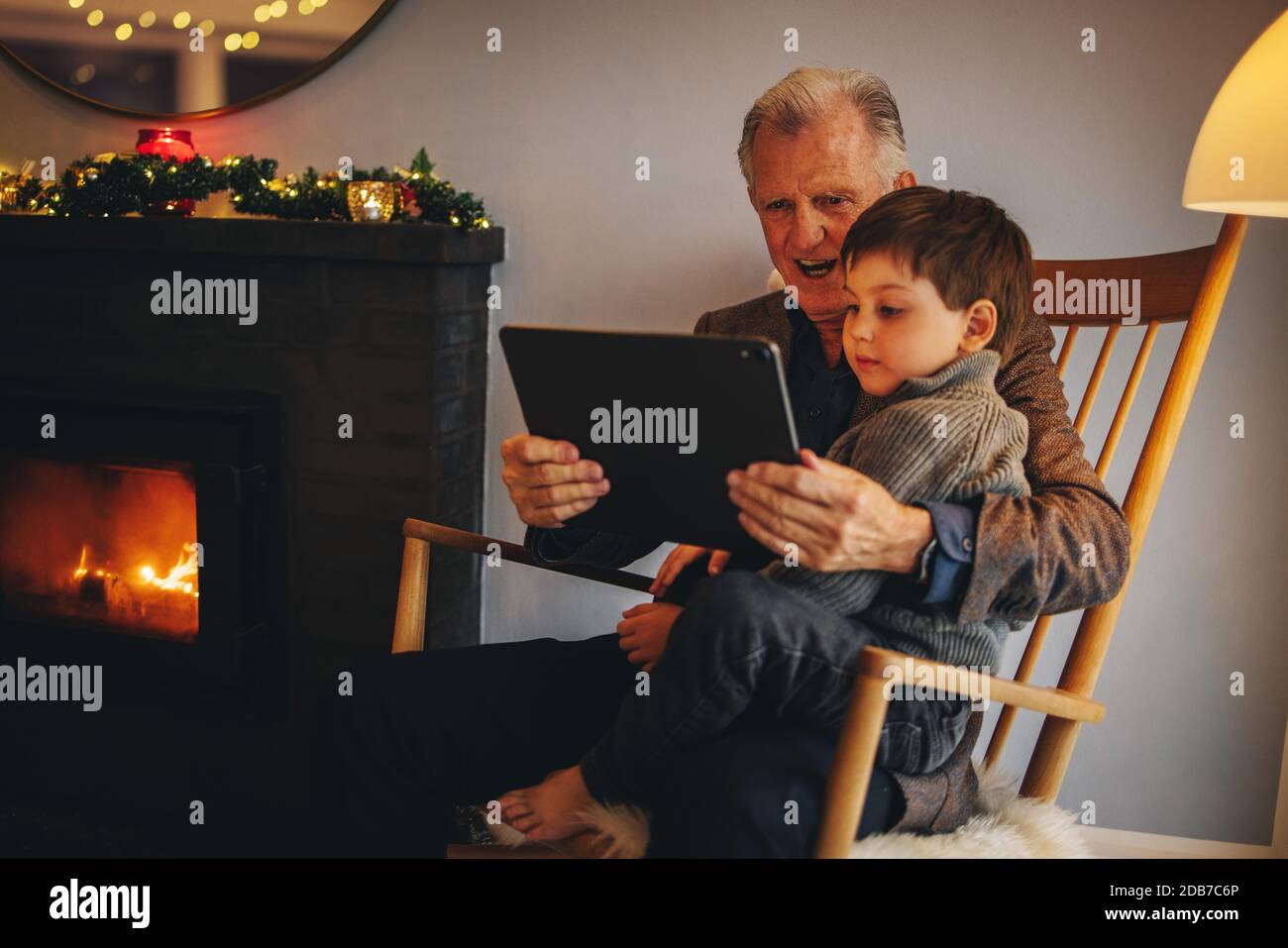 Älterer Mann sitzt auf einem Stuhl mit Enkel Blick auf digitale Tablet während weihnachten. Großvater und Enkel mit einem digitalen Tablet. Stockfoto