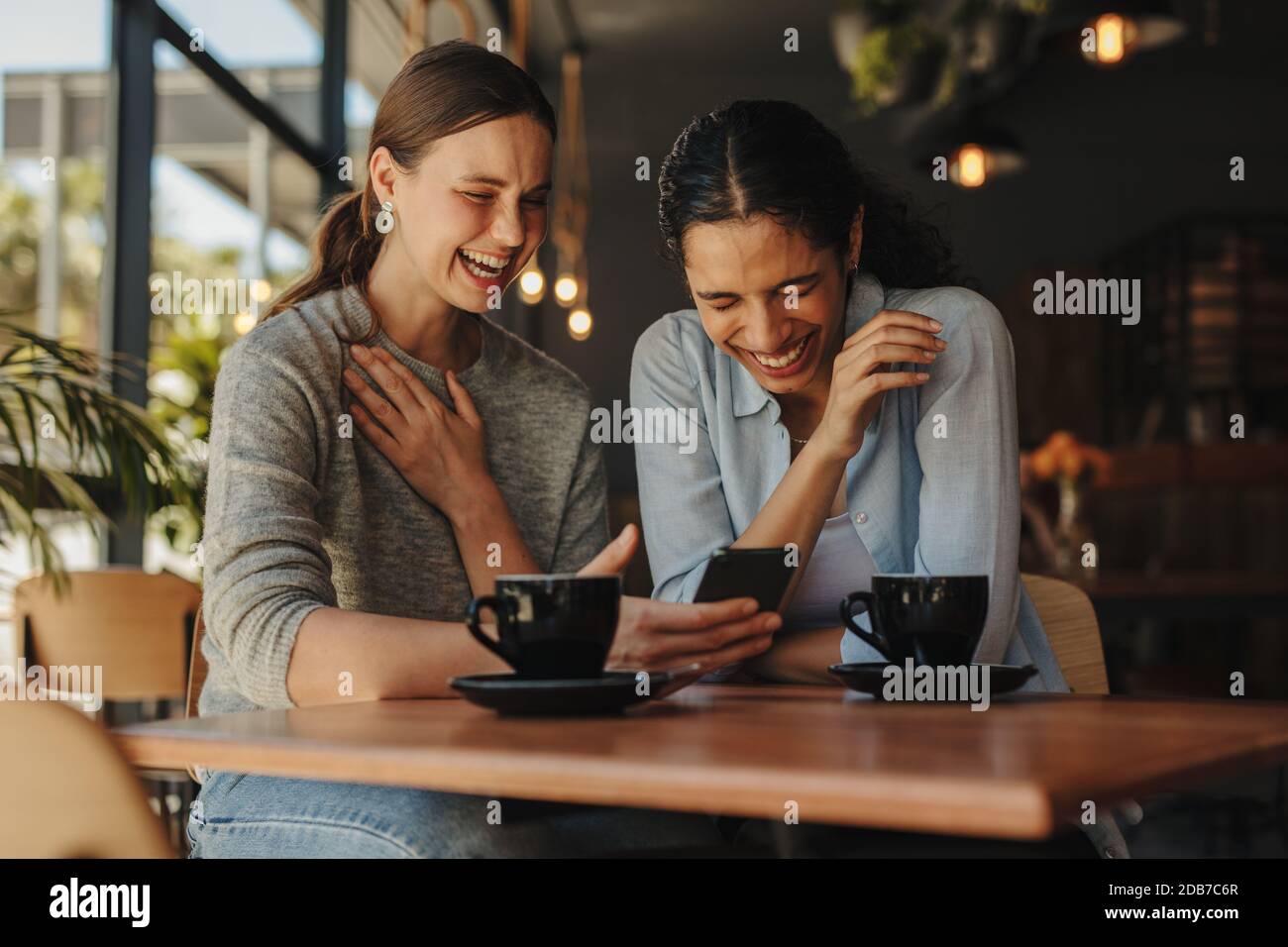 Zwei Freundinnen, die an einem Tisch im Café sitzen und auf ein Handy blicken und lächeln. Frau zeigt lustige Social-Media-Post auf ihrem Handy zu ihrem Fr. Stockfoto