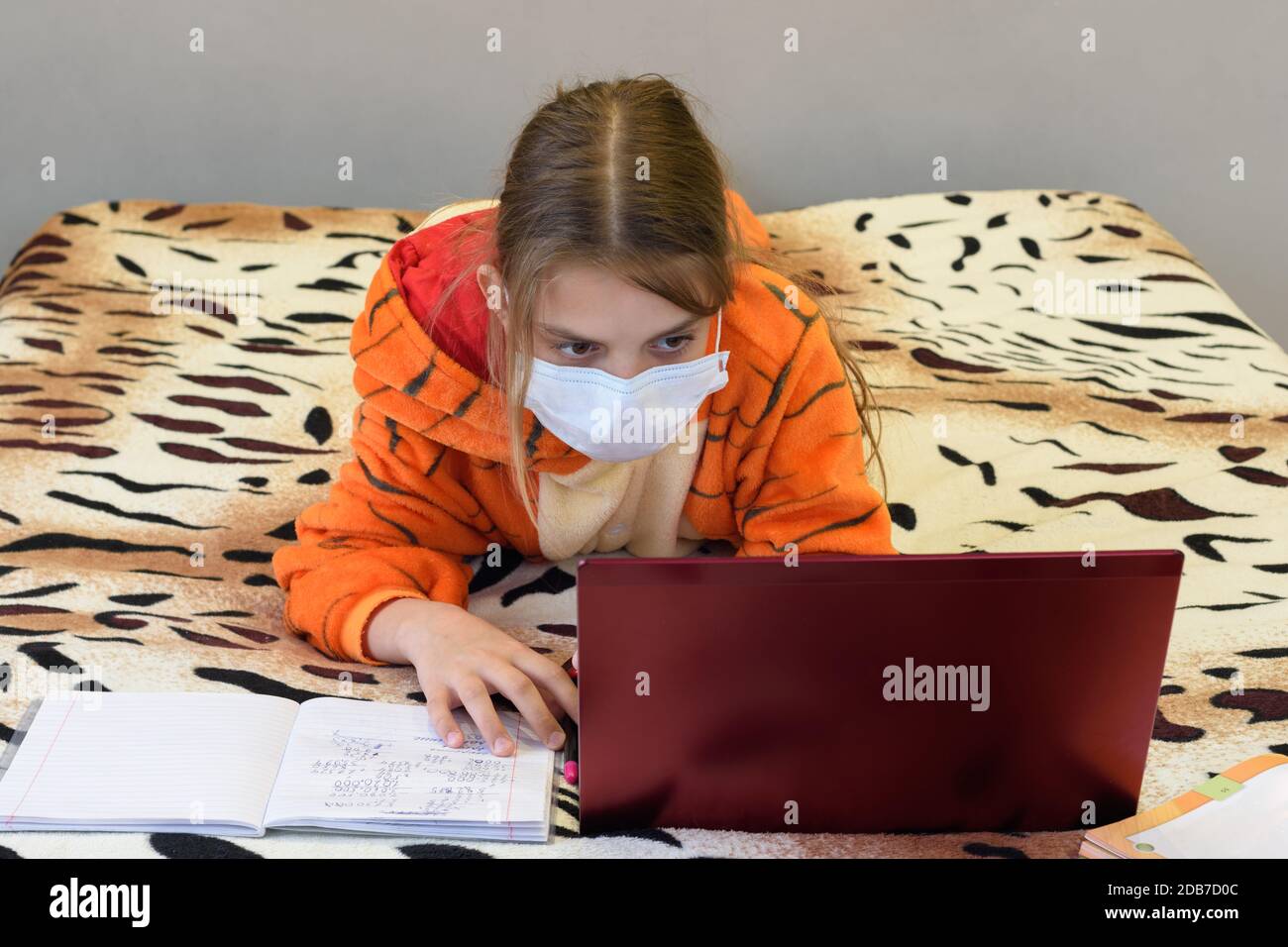 Ein Mädchen in einer medizinischen Maske im Schlafanzug liegt auf dem Bett und lernt ferngesteuert über das Internet Stockfoto