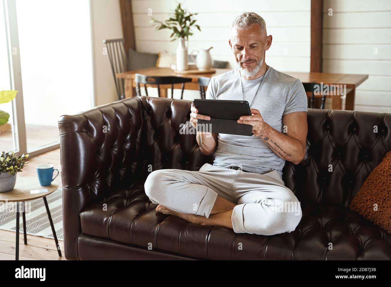 Kaukasischer Mann mittleren Alters in legerer Kleidung mit digitalem Tablet Während Sie im Wohnzimmer in Lotuspose auf dem Sofa sitzen Zimmer seiner modernen Wohnung Stockfoto