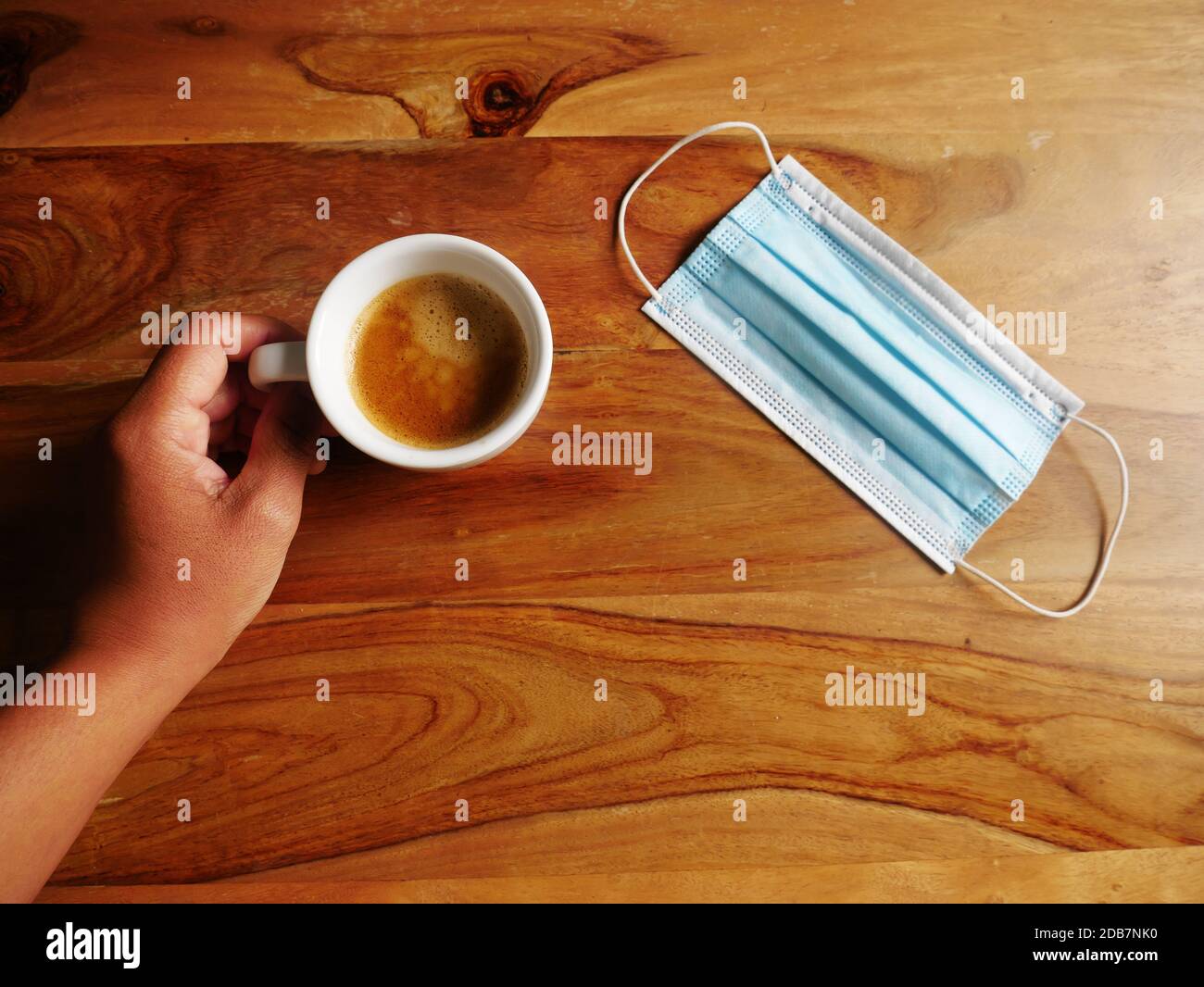 Foto einer Hand einer Person, die einen Kaffee nimmt und die ihre Maske auf dem Tisch hat. Stockfoto