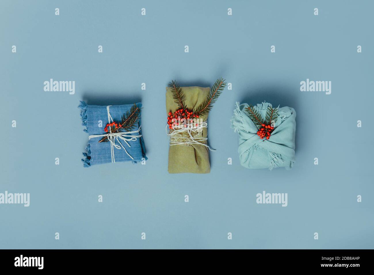 Draufsicht Zero Waste weihnachtskonzept. Drei Geschenke in Stoff eingewickelt mit Fichtenzweig und Beeren auf gedämpftem blauen Hintergrund. Flatlay. Minimalismus Stockfoto
