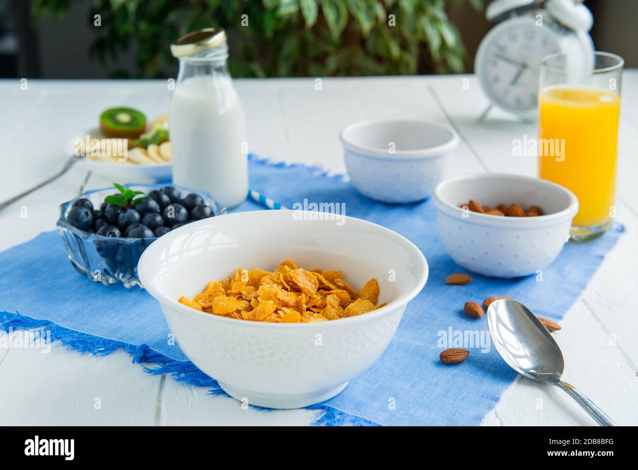 Nahrhaftes, gesundes Frühstück. Schüssel mit Getreide, Mandeln, Heidelbeere, Milch auf pflanzlicher Basis und Orangensaft auf blauer Serviette und weißem Holztisch Stockfoto