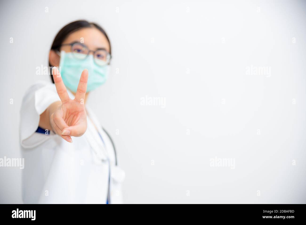 Studio Portrait schöne asiatische junge Frau Arzt heben zwei V-Form Finger tragen eine Maske zur Sicherheit Coronavirus im Kampf gegen Krankheit zu unterstützen Stockfoto