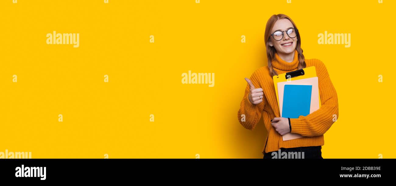 Schöne Schüler ist gestikulieren das wie Zeichen auf einem gelben studiowand lächelt die Kamera in der Nähe von freiem Raum und halten Ordner Stockfoto