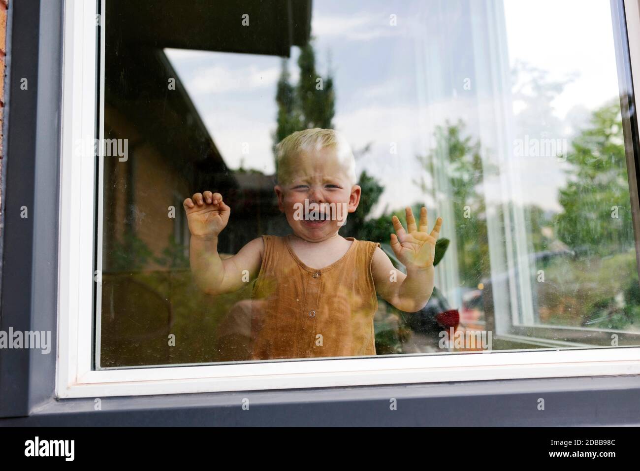 Kleiner Junge (18-23 Monate), der hinter dem Fenster steht und weint Stockfoto