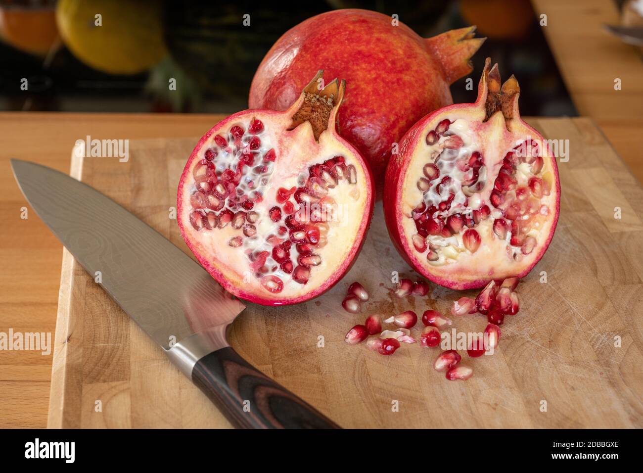 Nahaufnahme von einer ganzen und zwei halben Granatapfelfrüchten und einem damaszener Küchenmesser auf einem Holzschneidebrett mit Granatapfelkernen im Vordergrund Stockfoto
