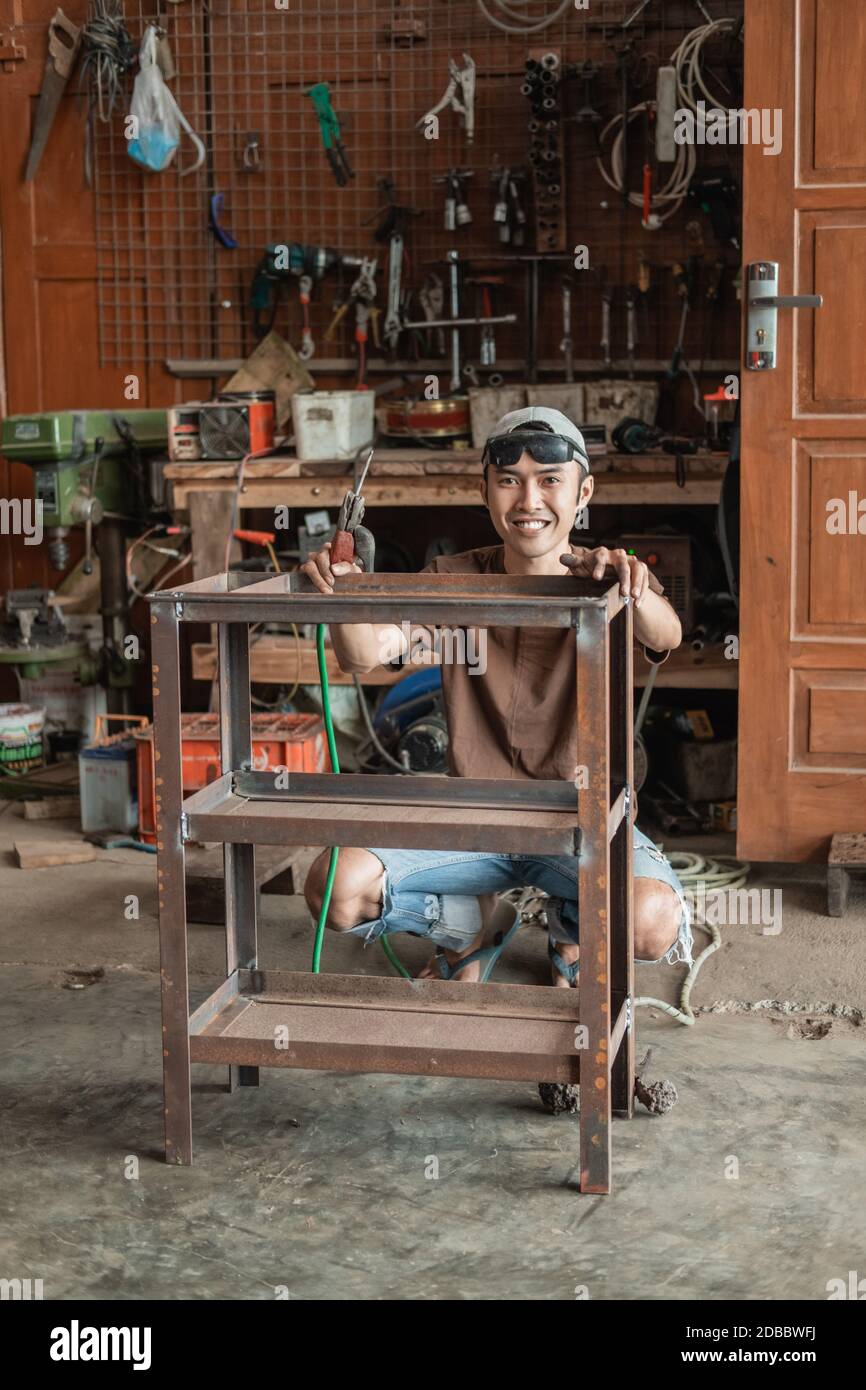 Squat männlich Schweißer lächelt auf die Kamera hält ein Eisen Rack zum Schweißen vor dem Hintergrund einer Schweißwerkstatt Stockfoto