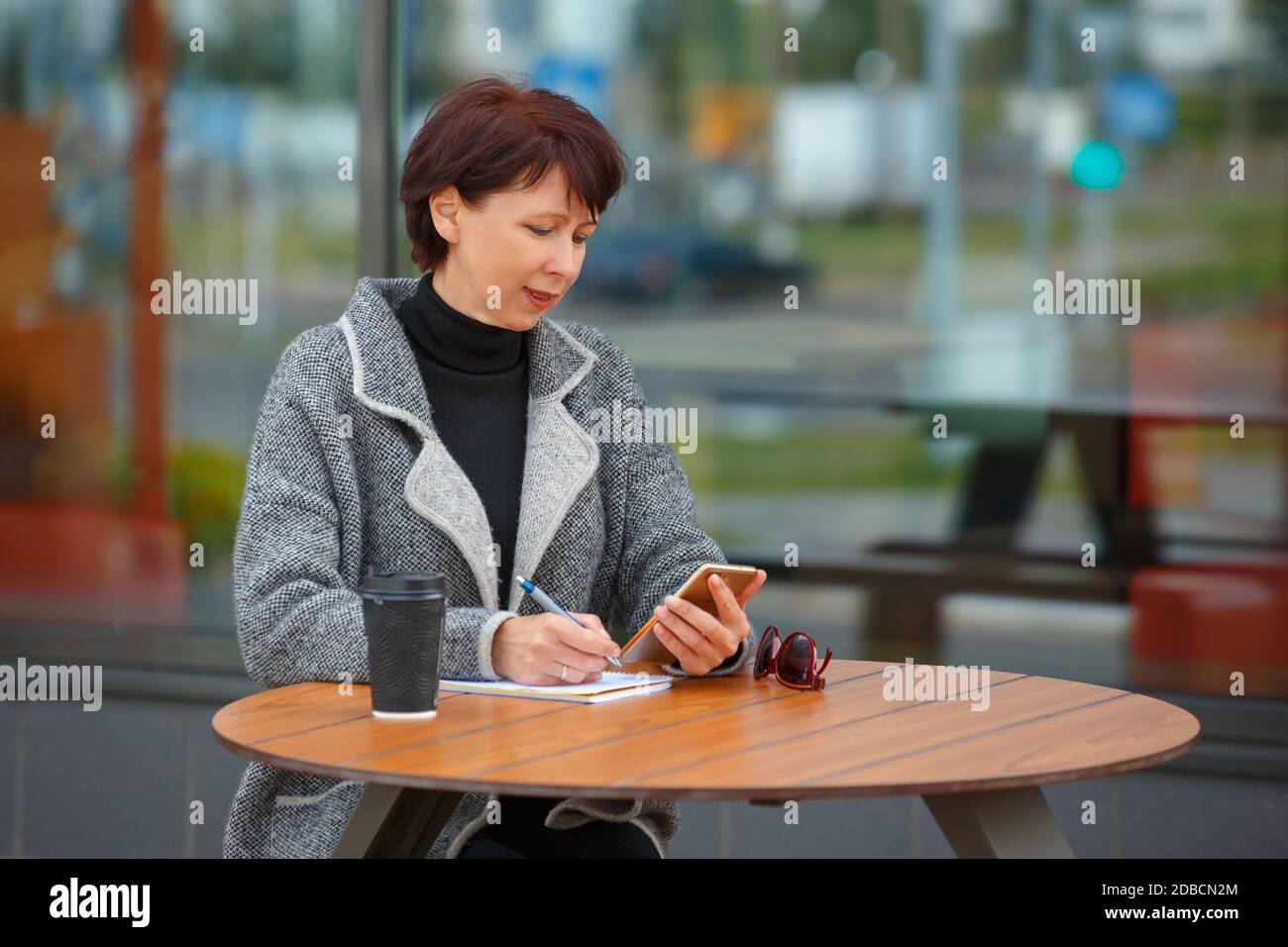 Eine Geschäftsfrau sitzt an einem Tisch in einem Sommercafé. Eine Frau hält ein Smartphone in der Hand und schreibt in ein Notebook. Stockfoto