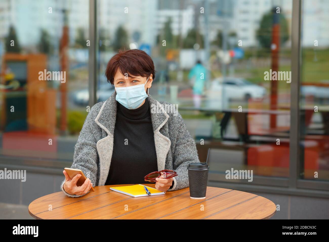 Eine Frau in einer Schutzmaske sitzt an einem Tisch in einem Sommercafé. Eine Frau hält ein Smartphone in der Hand und schreibt in ein Notebook. Stockfoto