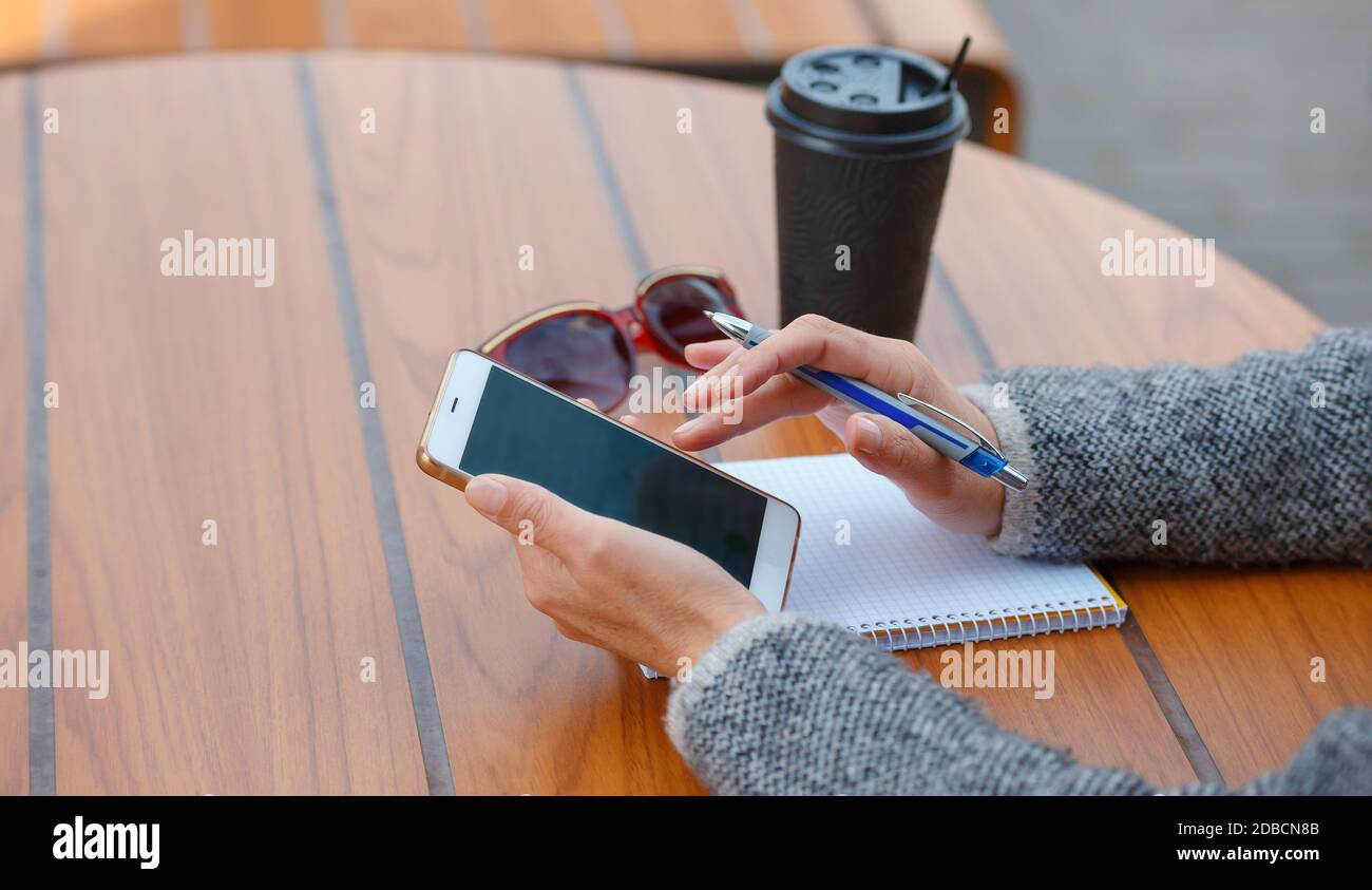 Eine Geschäftsfrau sitzt an einem Tisch in einem Sommercafé. Eine Frau hält ein Smartphone in der Hand und schreibt in ein Notebook. Stockfoto