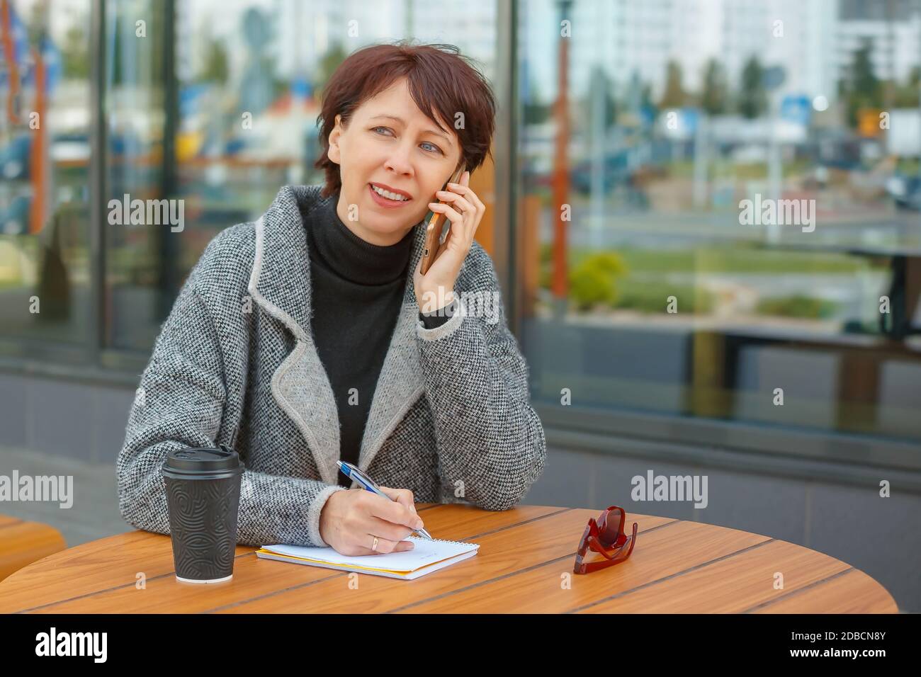Eine Geschäftsfrau sitzt an einem Tisch in einem Sommercafé. Die Frau spricht auf dem Smartphone und schreibt in einem Notebook. Stockfoto