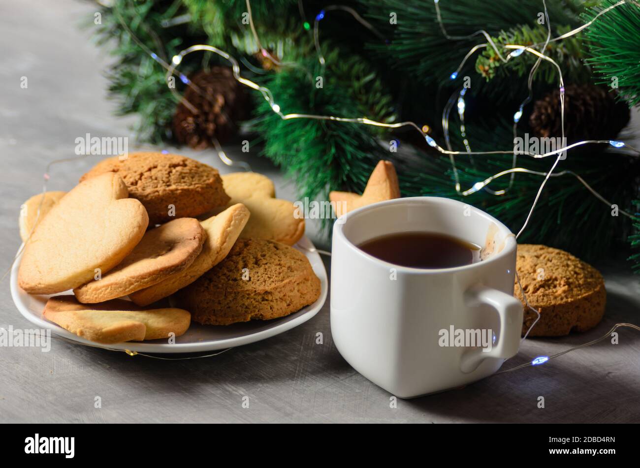 Hausgemachte Kekse in Form eines Herzens auf einem weißen Teller mit einem Becher und einem Teebeutel auf dem Hintergrund der Weihnachtsdekoration. Selektiver Fokus. Stockfoto