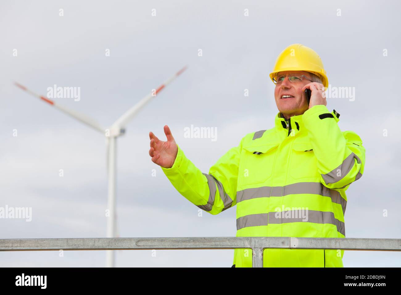 Techniker mit Schutzarbeiten tragen Gespräch auf Mobiltelefon in Vorderseite einer Windenergieanlage im unscharfen Hintergrund Stockfoto