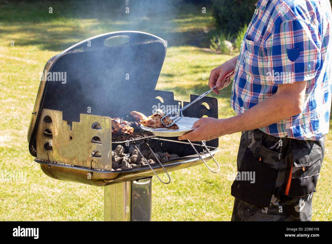 Ein Mann am Holzkohlegrill grillen Steaks, Grillwürste und Hühnerflügel und  benutzen Grillzangen. Selektiver Fokus auf den Grill gegen eine gree  Stockfotografie - Alamy