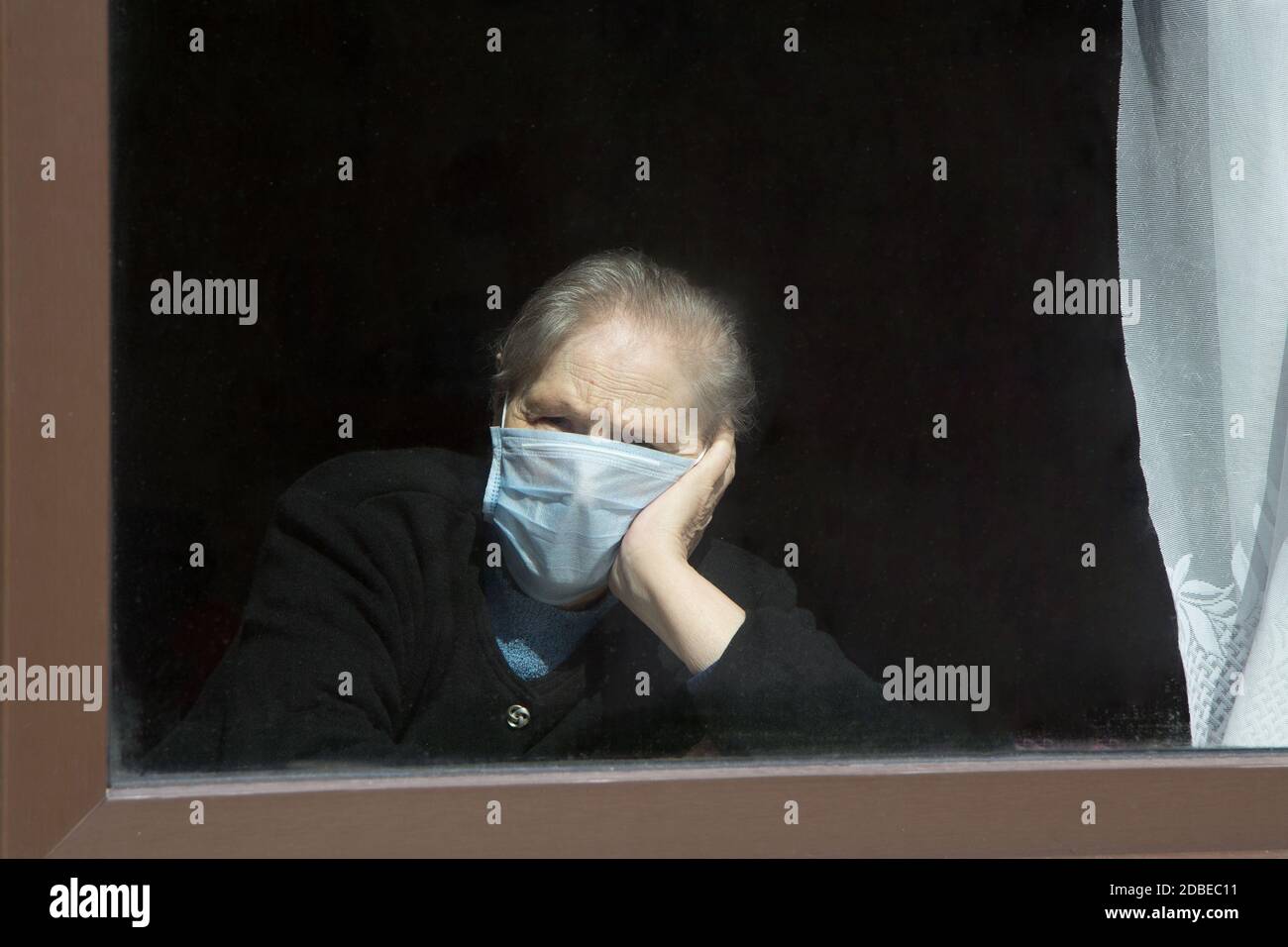 Eine alte Großmutter in einer Schutzmaske blickt aus dem Fenster zur Selbstisolierung. Ältere Frau in Quarantäne zu Hause. Stockfoto