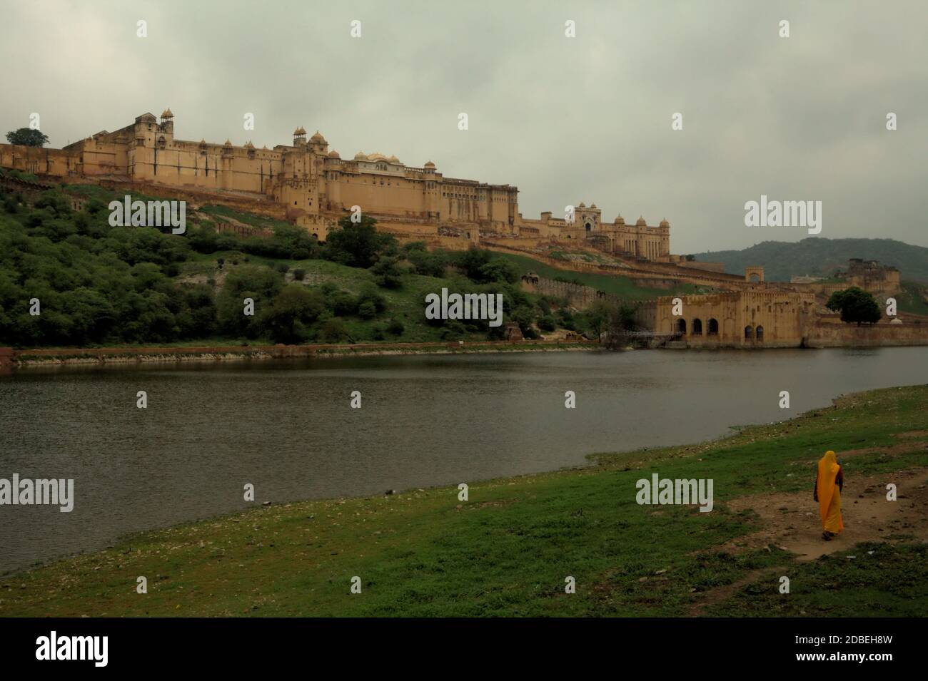 Eine Frau in Arbeitskleidung zu Fuß auf der Seite des Maota Lake, mit Blick auf die historische Amer Fort in Amer, Rajasthan, Indien. Stockfoto