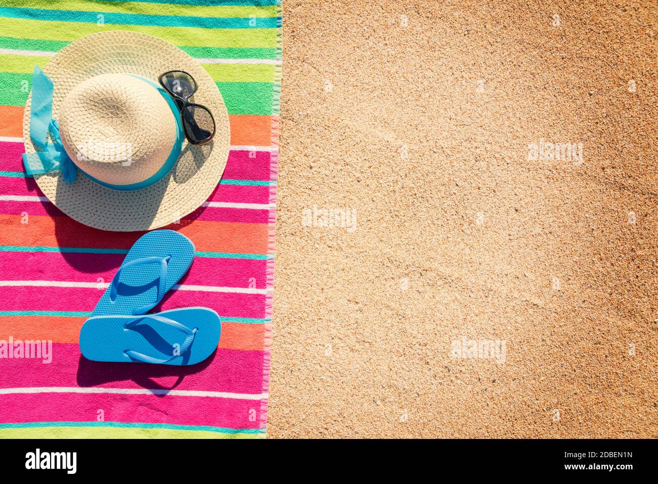 Strandtuch mit Hut, Sonnenbrille und Flip Flops von oben am Sandstrand fotografiert, heiße Sommer-Tages-Accessoires, Urlaubsziel, sonniger tropischer Strand Hintergrund Stockfoto