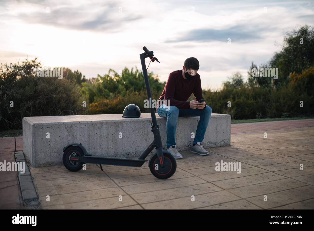 Junger Mann auf einem Elektro-Skateboard mit einem Handy sitzt auf einer Bank mit seinem Helm. Alternativer Transport. Stockfoto