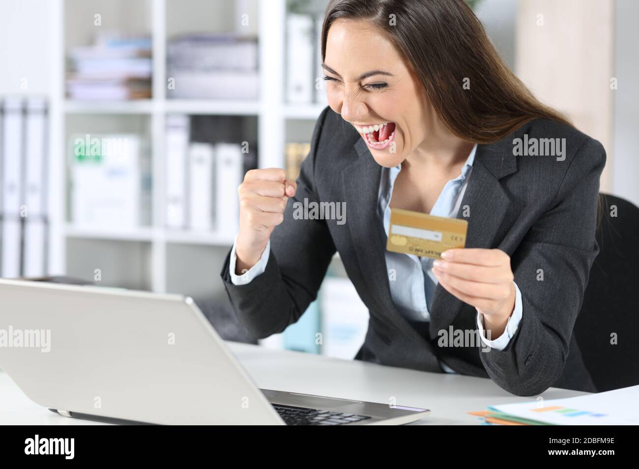 Aufgeregt Executive Frau online mit Kreditkarte auf Laptop zahlen sitzt auf einem Schreibtisch im Büro Stockfoto