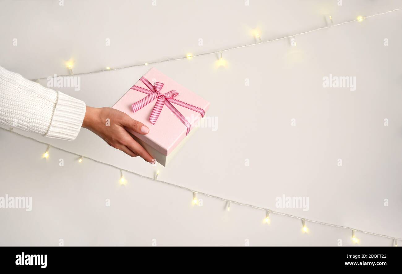 Rosa Geschenkbox mit Schleife in weiblicher Hand in weißem Pullover auf hellem Hintergrund. Weihnachten Girlanden an der Wand Stockfoto