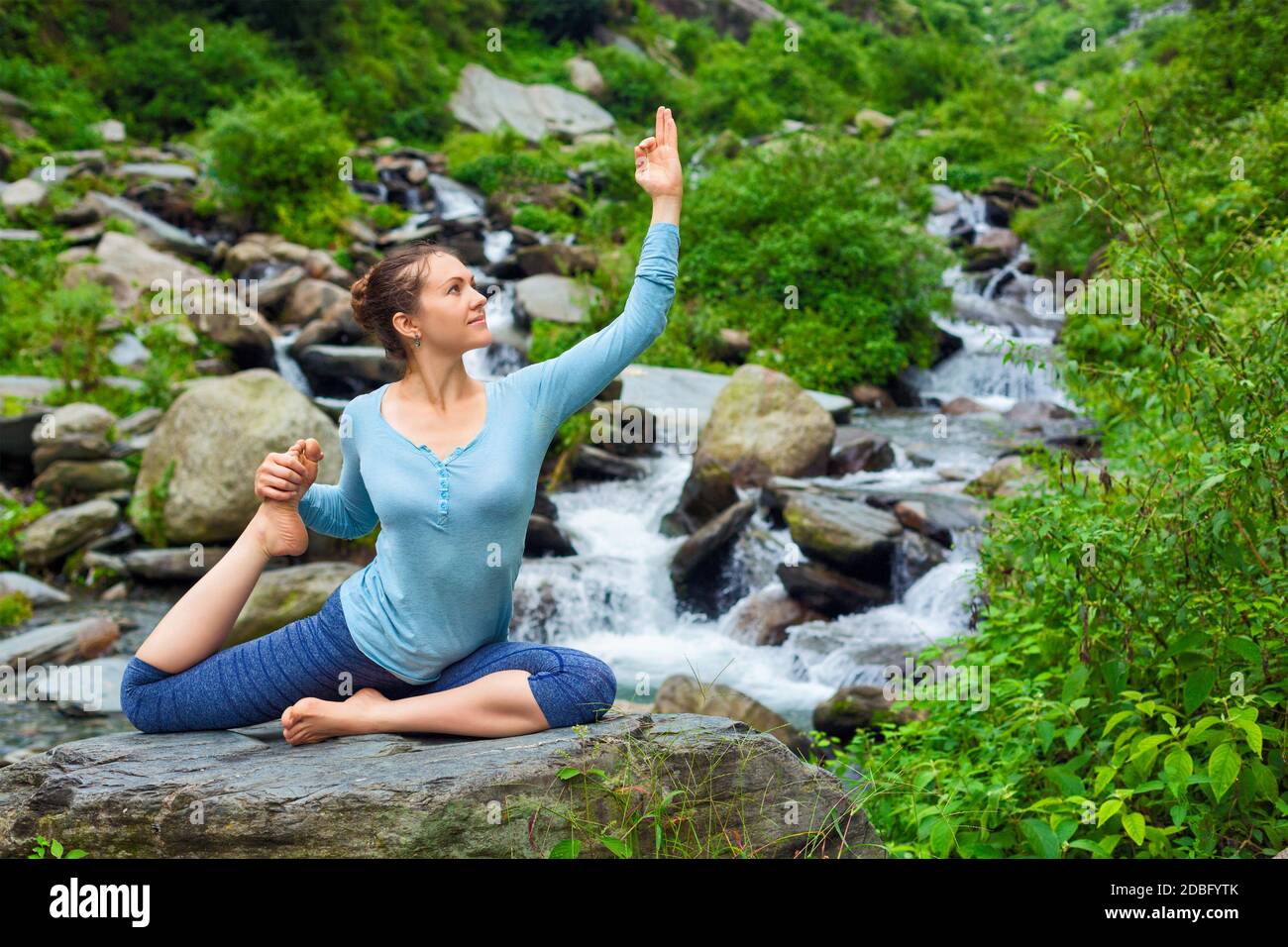 Hatha Yoga Outdoor - junge sportliche fit Frau macht Yoga asana Eka pada rajakapotasana - einbeinige Königstaube Pose am tropischen Wasserfall. Himachal Pr Stockfoto