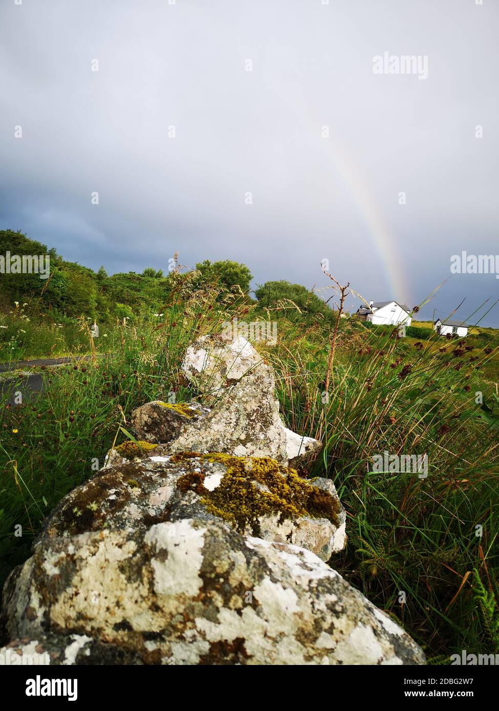 Irische Landschaft von Connemara mit wildem Gras, das an der Seite der alten Steinmauer wächst, kleines Häuschen in der Ferne und hellem Regenbogen Stockfoto