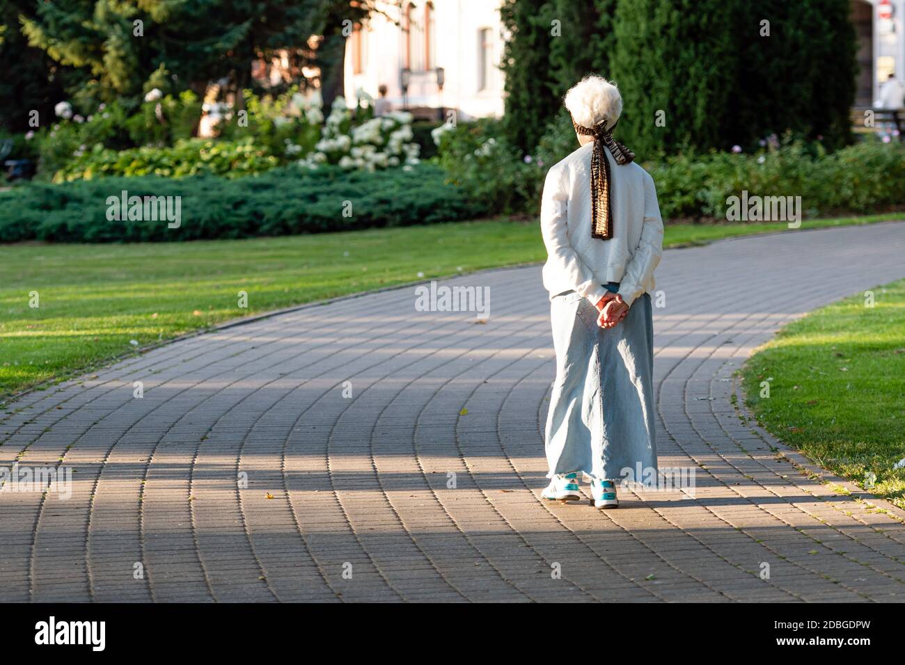 Eine ältere Frau mit grauen Haaren und einem langen Rock Geht weg auf dem gepflasterten Gehweg des Parks Stockfoto