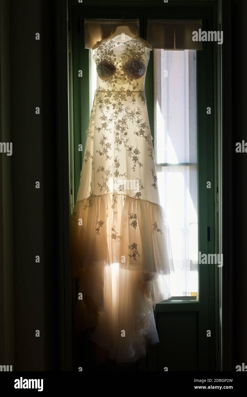 Hintergrundbeleuchtetes elegantes Hochzeitskleid, das in einem Fenster hängt und das zeigt Zartes Muster des Stoffes in einem abgedunkelten Raum Stockfoto