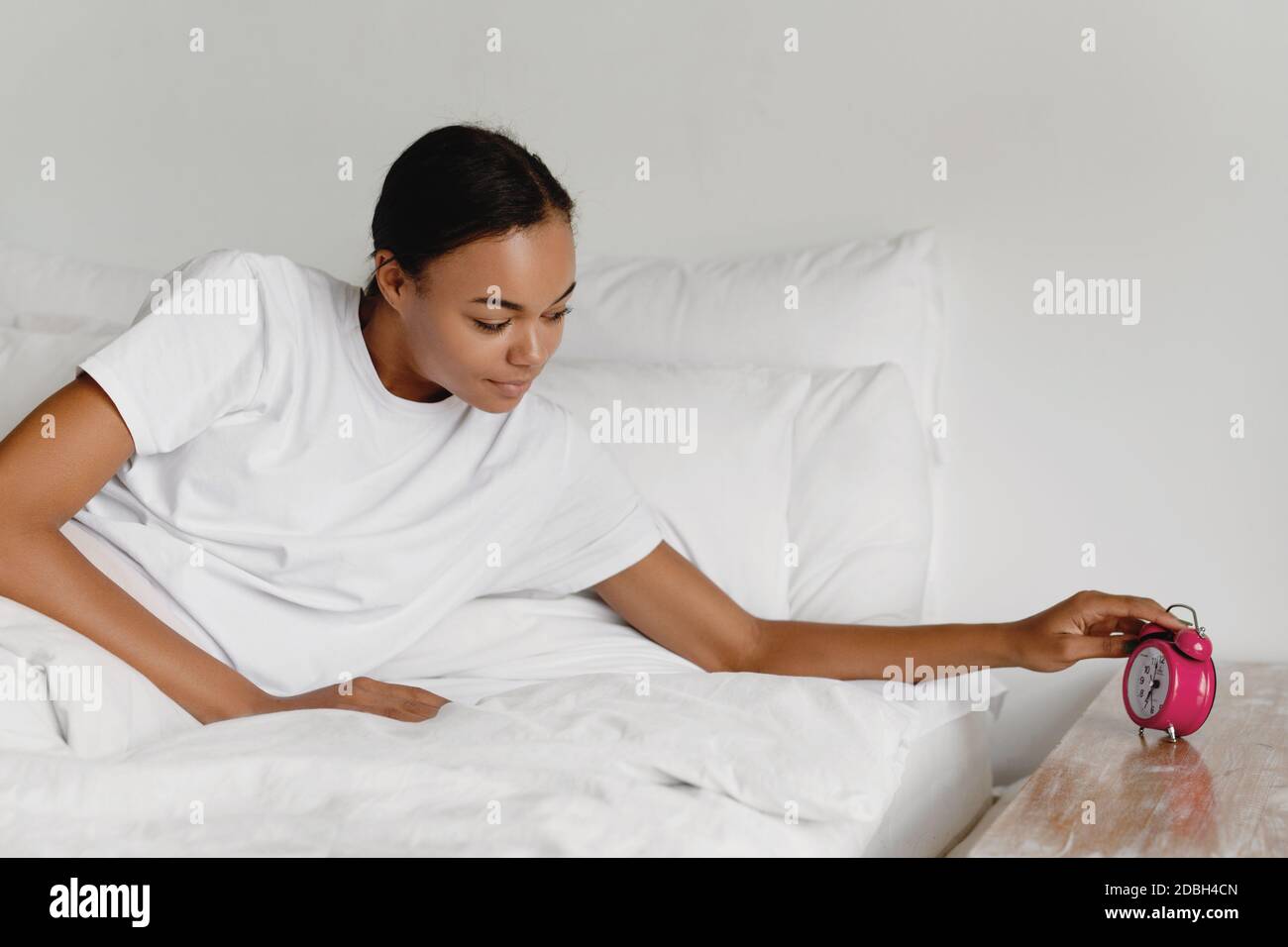 Junge afroamerikanische Frau, die aufwacht und die Hand zum rosa Wecker reicht. Stockfoto
