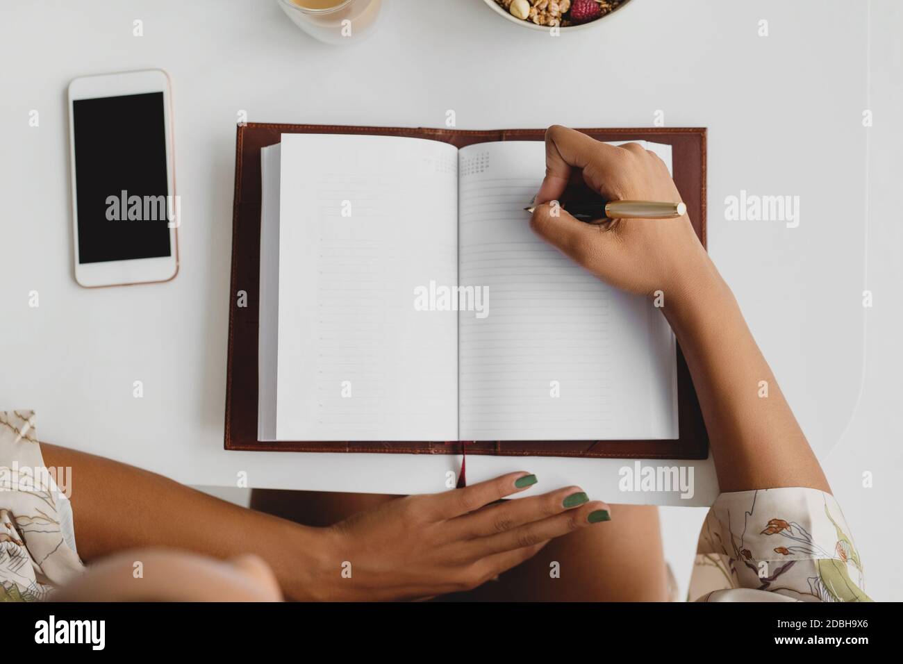 Schwarze weibliche Hände, die beim Frühstück Notizen im Notizbuch auf weißem Tischhintergrund machen. Draufsicht. Stockfoto