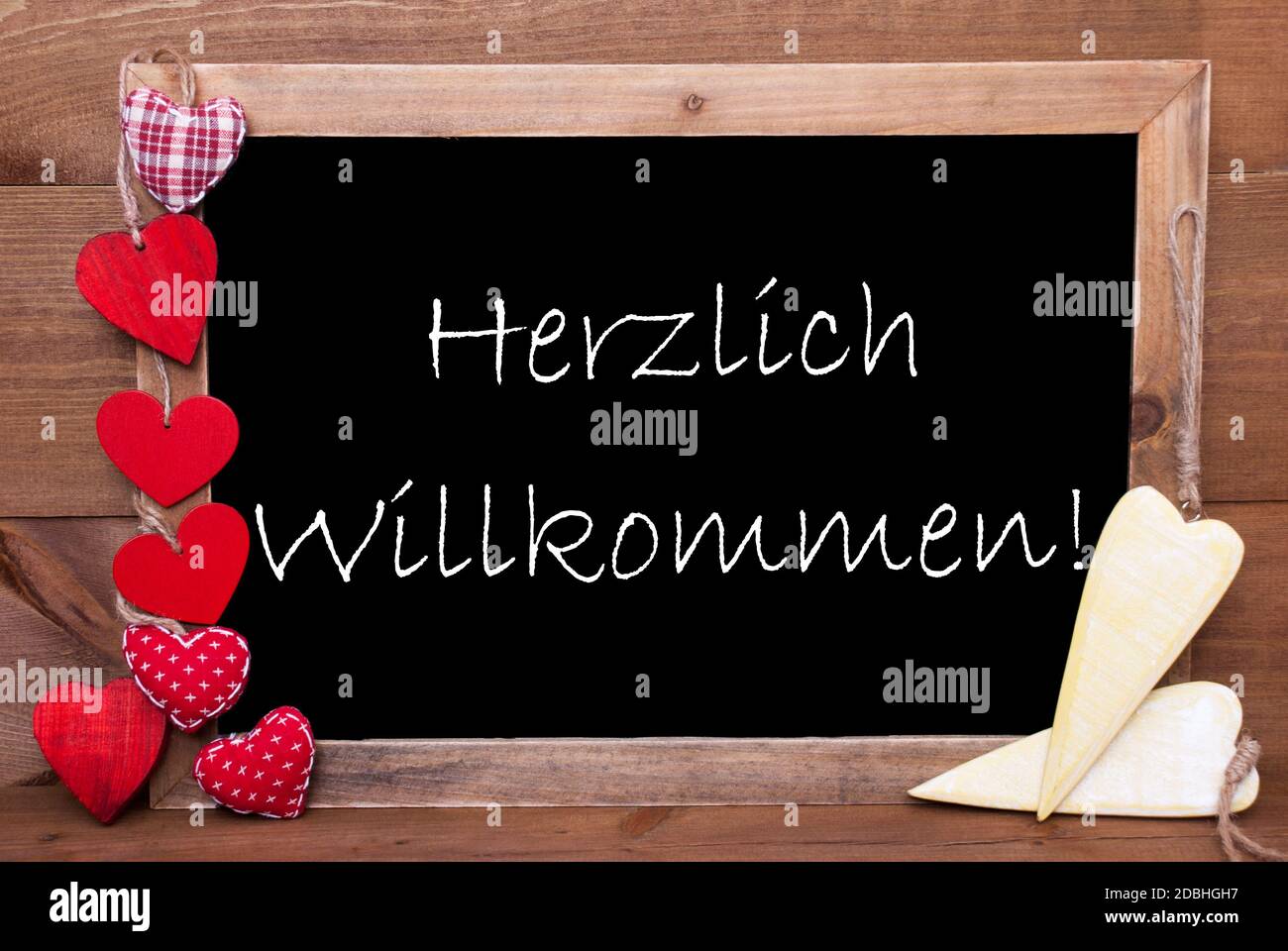 Balckboard Mit Deutschem Text Herzlich Willkommen Heißt Willkommen. Rote Und Gelbe Herzdekoration. Brauner Holzhintergrund Stockfoto