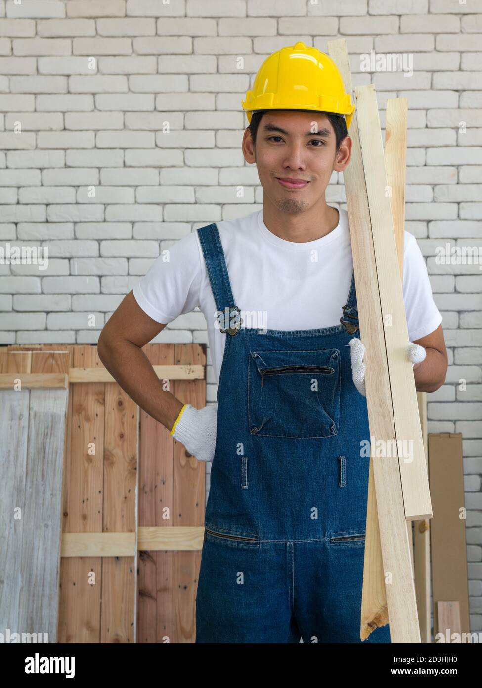 Ein asiatischer Zimmermann trägt einen gelben Hardhat und trägt Holz für die Möbelherstellung im Werkstattraum. Stockfoto