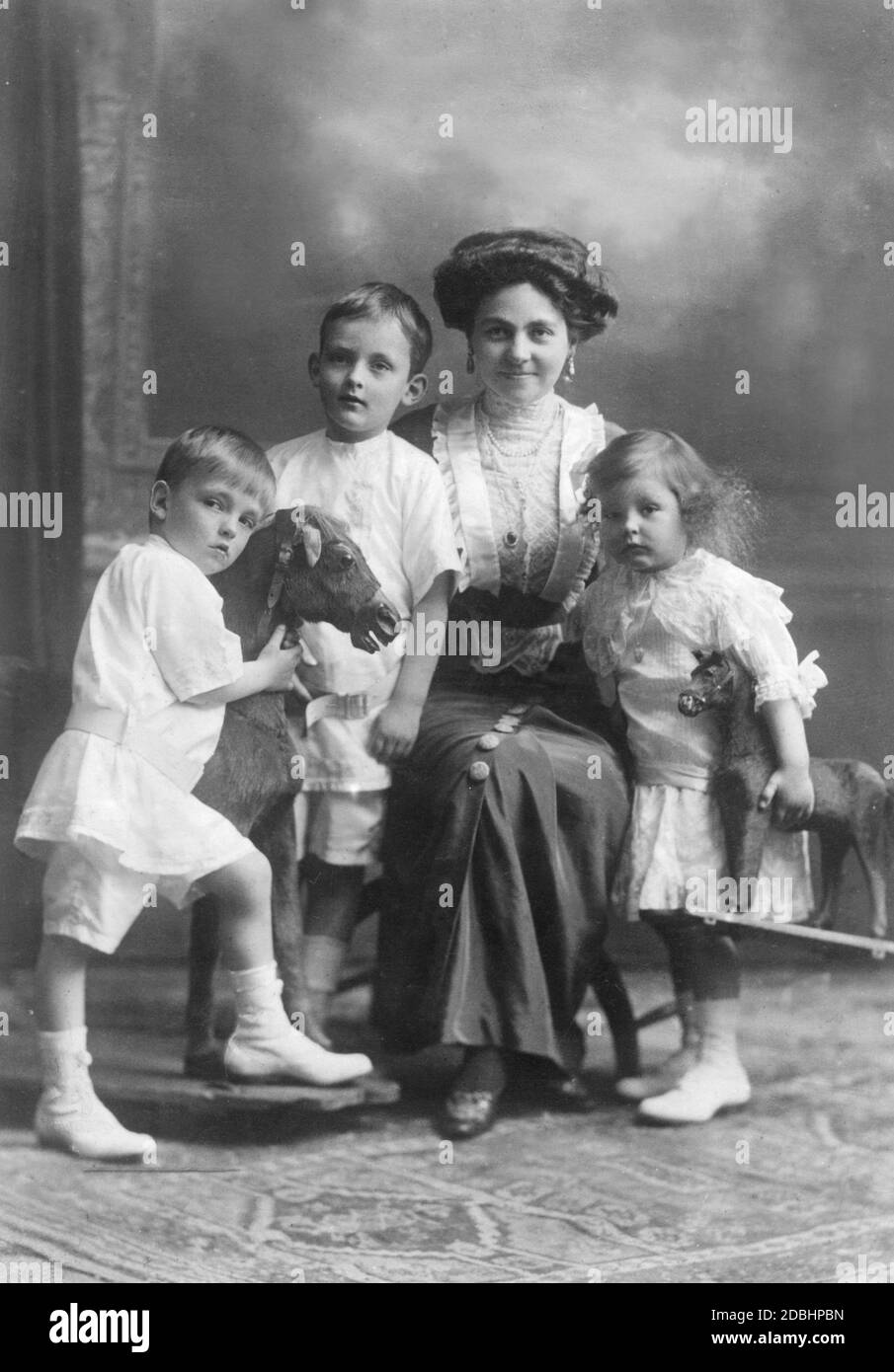 Hermine Reuss Elder Line mit ihren drei ältesten Kindern, von links nach rechts: Prinz Georg Wilhelm von Schoenaich-Carolath, Prinz Hans Georg von Schoenaich-Carolath, Hermine Reuss Elder Line und Prinzessin Hermine Karoline von Schoenaich-Carolath. Die Kinder haben ein Schaukelpferd und ein weiteres Spielzeugpferd zum Spielen. Das Bild wurde um 1913 aufgenommen. Stockfoto