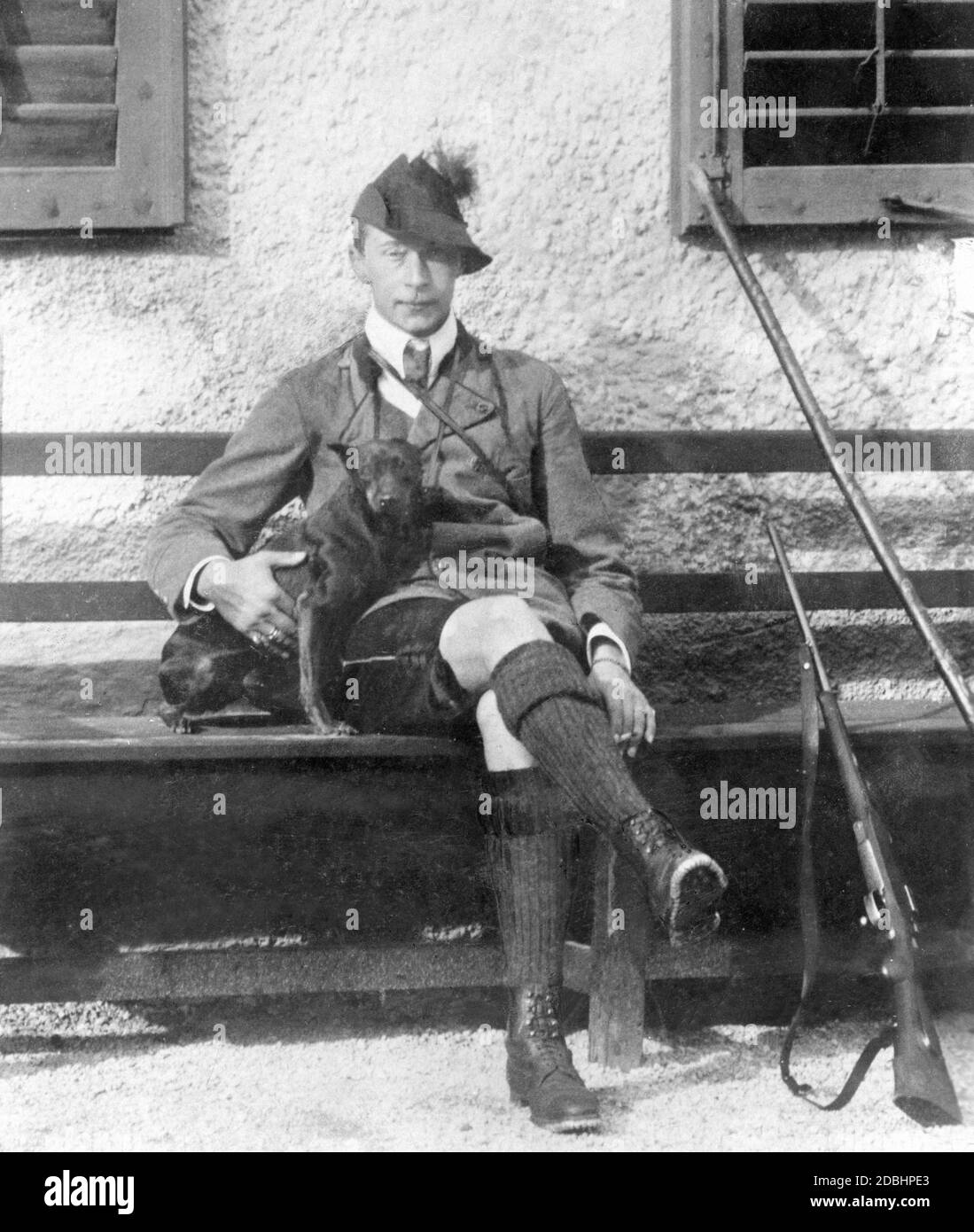 Kronprinz Wilhelm von Preußen, in bayerischer Tracht, sitzt mit seinem Hund Tricky und einem Jagdgewehr neben ihm 1906 vor einem Haus in Wildbad Kreuth. Er war Jagdgast von Herzog Karl Theodor von Bayern. Stockfoto