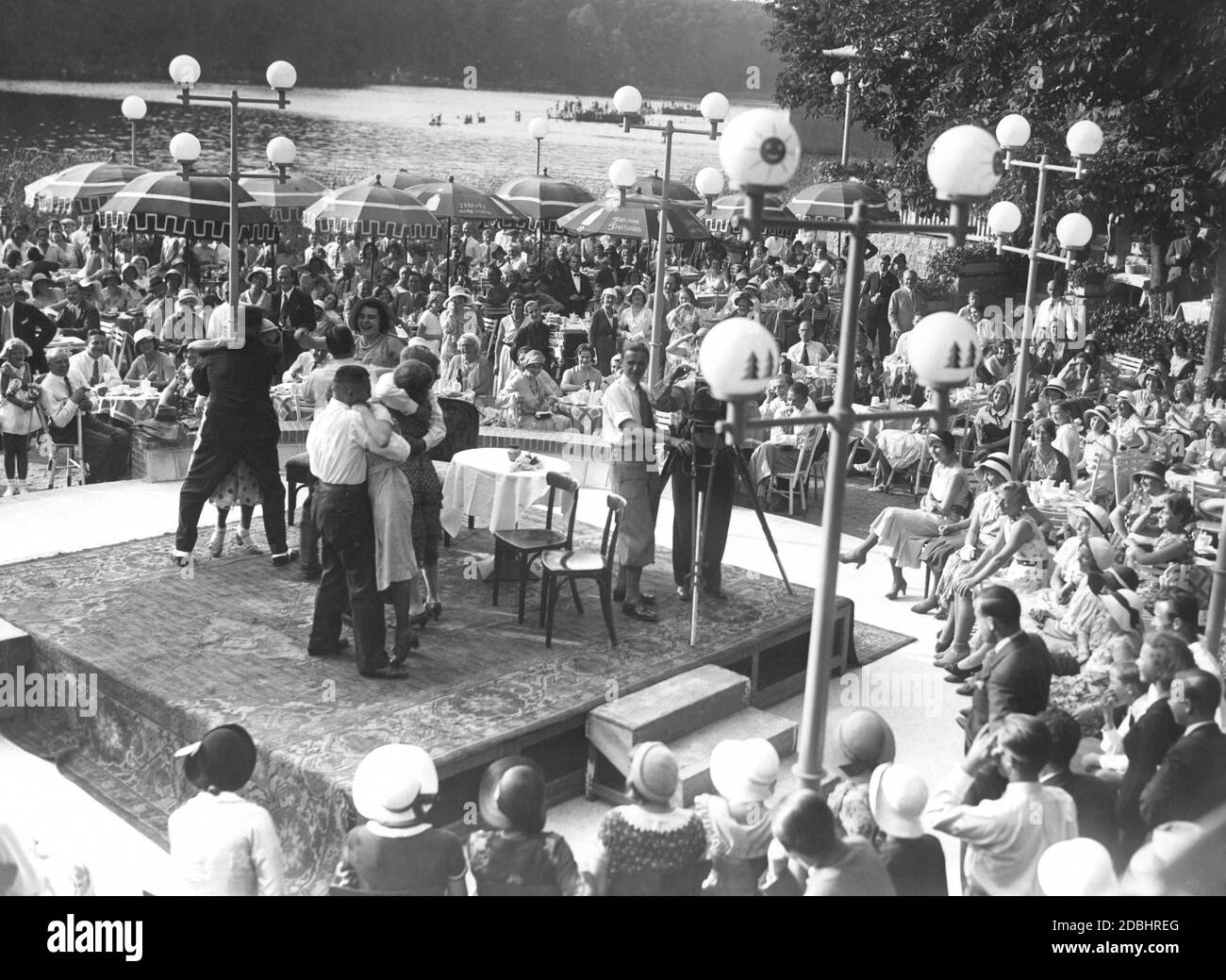 "An einem schönen Sommertag 1931 fand am Krumme Lanke See in Berlin-Zehlendorf ein Filmfestival statt. Das Publikum spielt und ihre Emotionen und Reaktionen werden von einer Tonfilmkamera aufgenommen, die auf der Bühne steht. Auf zwei Sonnenschirmen wird Milch mit den Slogans 'Milch macht stark' und 'frisch durch frische Milch' beworben. Auf der Bühne steht eine Kamera, die auf die Kamera des Fotografen zeigt." Stockfoto