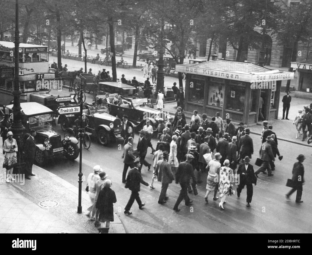 1929 überqueren Fußgänger die Straße unter den Linden in Berlin an der Kreuzung mit der Friedrichstraße. Auf der rechten Seite ist ein kleines Haus mit Informationen über Bad Kissingen. Ein Doppeldeckerbus der Linie 11 fährt zum Hermann-Platz in Neukölln. Stockfoto