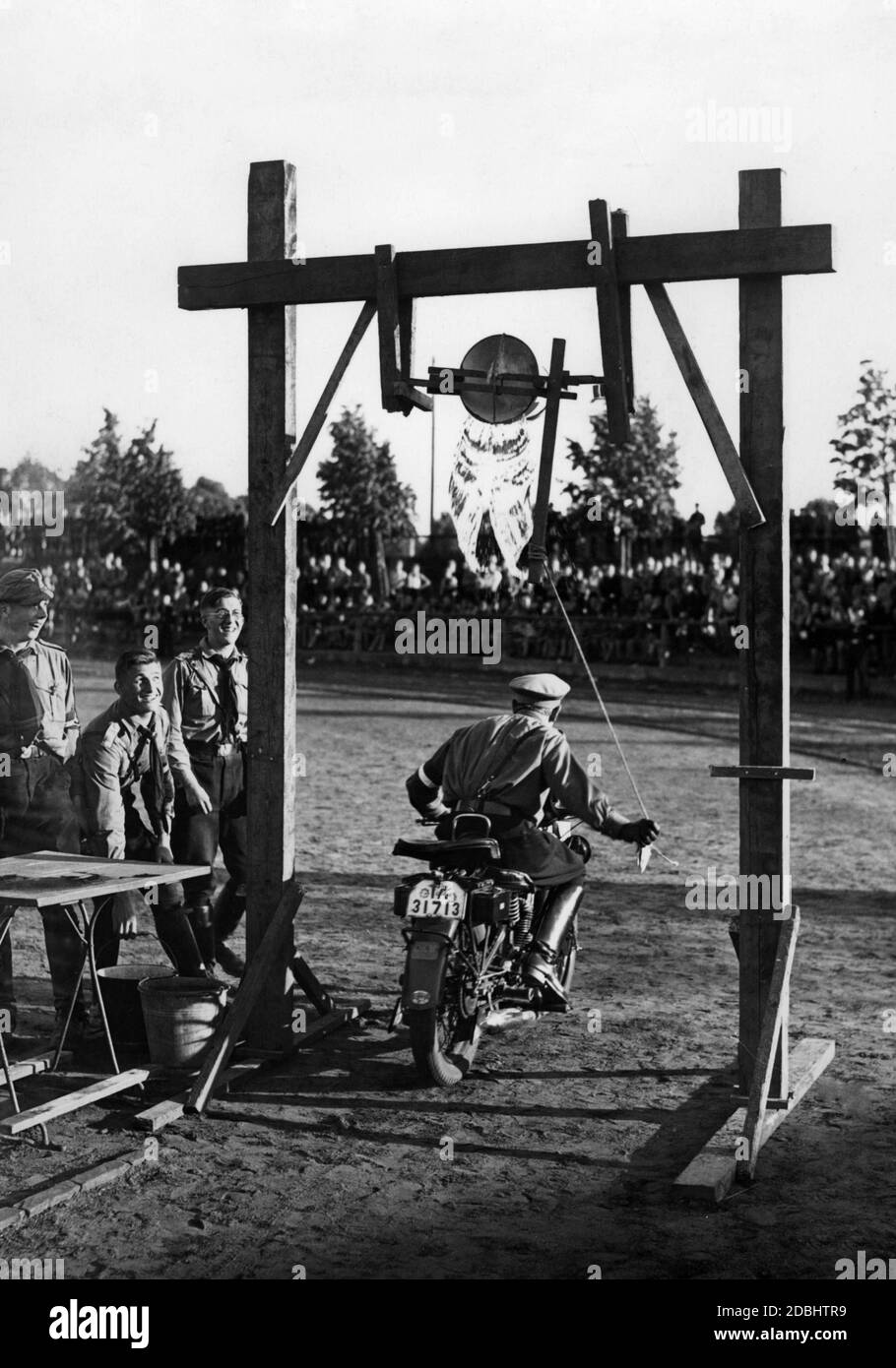 Ein Mitglied der Motor HJ bei einem Fahrtest während des Sportfestivals der Hitlerjugend auf dem Westend Sportplatz in Berlin. Er fährt unter einem Wassereimer. Das Publikum ist im Hintergrund. Auf der linken Seite des Bildes sind andere Mitglieder des HJ lachend. Er fährt ein Motorrad der Marke DKW. Stockfoto