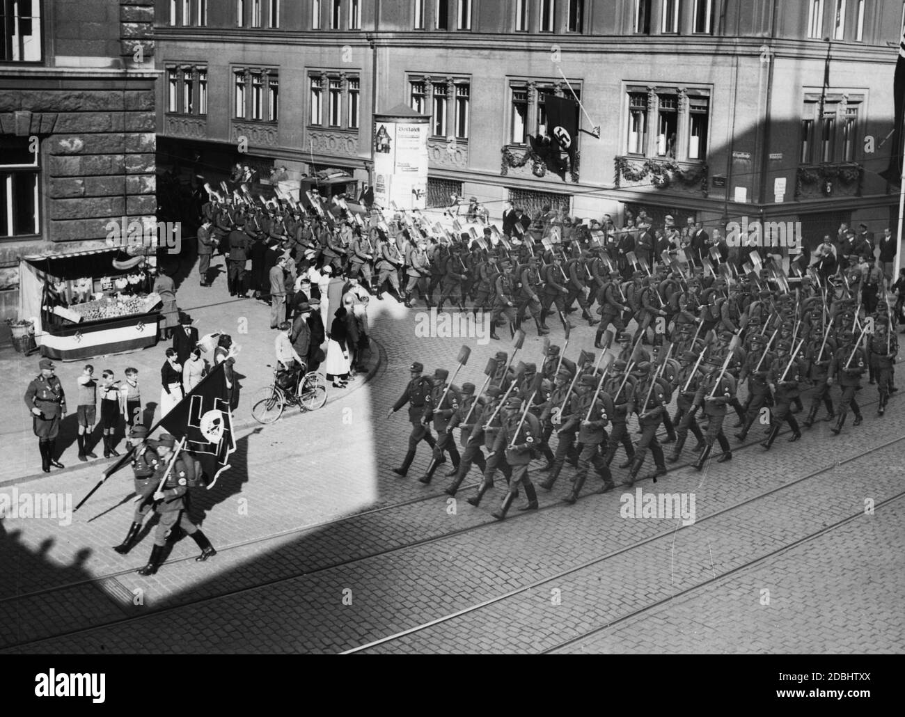 Mitglieder des Reichsarbeitsdienstes marschieren hinter ihrer Flagge mit Pik in den Händen zu ihrem Quartier auf dem Reichsparteitagsgelände. Am Straßenrand werden sie von der Bevölkerung begrüßt. Auf der linken Seite ist ein Obststand mit der Reichsflagge und Hakenkreuzen geschmückt. Stockfoto