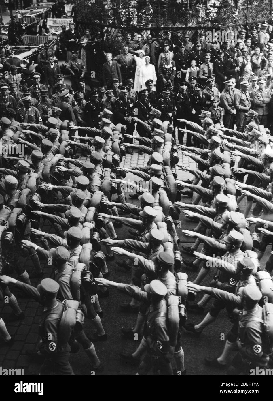 SA-Einheiten marschieren während des NSDAP-Kongresses durch Nürnberg. Sie werden von der Bevölkerung und von SS-Führern beobachtet, die in der Mitte lächeln. Links im Hintergrund sind mehrere Mercedes-Autos zu sehen. Stockfoto