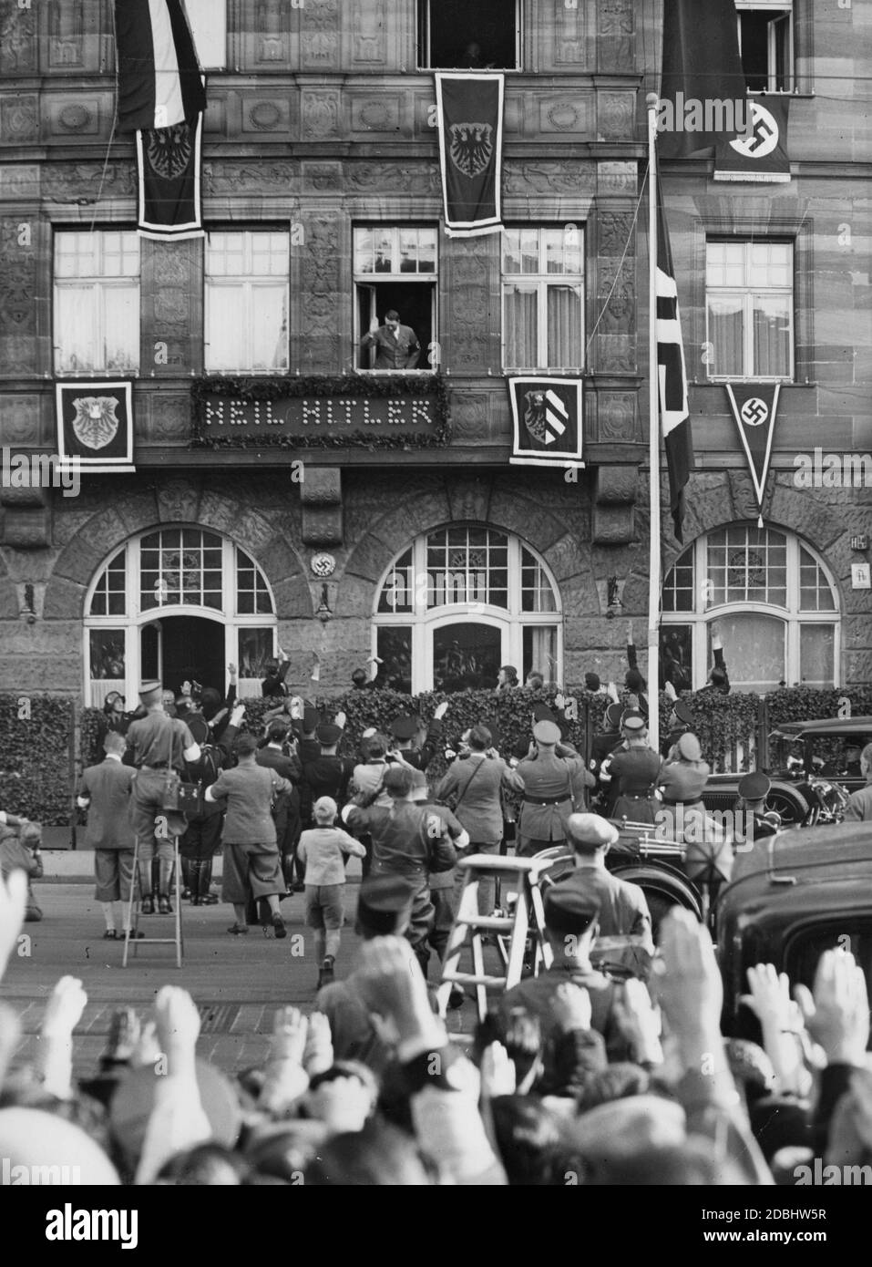 "Adolf Hitler begrüßt die begeisterte Nürnberger Bevölkerung aus dem Fenster des Hotels Deutscher Hof, das mit Hakenkreuzfahnen und dem Wappen der Stadt Nürnberg geschmückt ist. Unter seinem Fenster ist die Inschrift ''Heil Hitler''.' Stockfoto