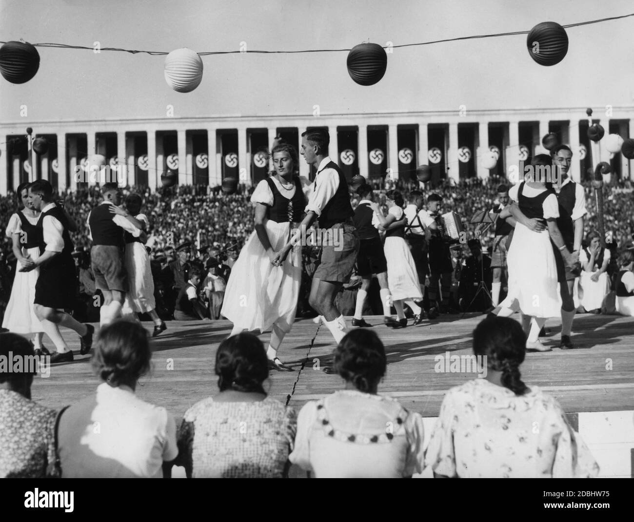 Eine Volkstanzgruppe in traditionellen Kostümen tanzt auf einem erhöhten Podium während des Volksfestes auf dem Zeppelin-Feld. Im Hintergrund die Zeppelin Tribüne. Stockfoto