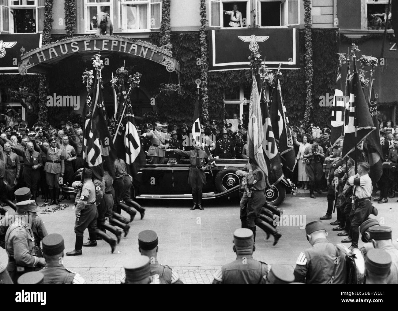 SA-Mitglieder marschieren mit ihren Sturmfahnen an Adolf Hitler vorbei. Rudolf Hess steht vor dem Auto. Hinter Adolf Hitlers Auto steht die Blutfahne. Im Hintergrund ist der Eingang zum Haus Elephant. Stockfoto