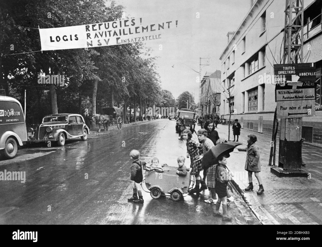 "Kinder mit einem Kinderwagen stehen an der Seite einer Straße in Beauvais. Über der Straße hängt ein Banner mit der Aufschrift "Refugies! Logis ravitaillement! NSV-Betriebsstandort''. Die National Socialist People's Welfare Organization kümmert sich um französische Flüchtlinge." Stockfoto