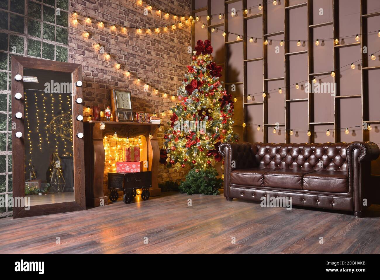 Stilvolle Weihnachts-Loft-Dekorationen in einem braunen Wohnzimmer mit einem Ledersofa, Weihnachtsbaum und Girlanden mit Glühbirnen an der Wand Stockfoto