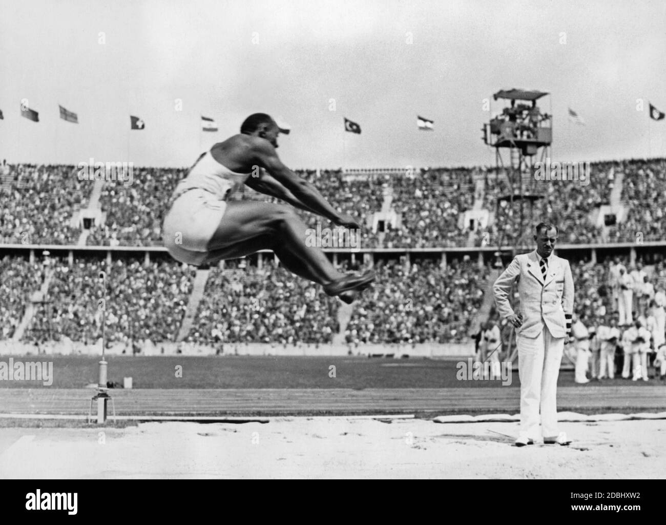 Jesse Owens bei einem Weitsprung-Wettbewerb. Im Hintergrund das Berliner Olympiastadion mit den Flaggen der teilnehmenden Nationen. Owens gewann die Goldmedaille in der olympischen Disziplin Weitsprung mit einem weltbesten Sprung von 8.06 m. Stockfoto